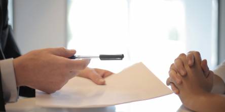 Two people sitting across from each other a desk, only their hands are visable. The one on the left is holding a contract and a pen.