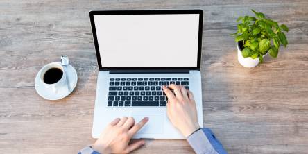 Macbook open on a desk with a black coffee to the left and a small green plant to the right. A man has both hands out and is typing on on the laptop.