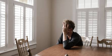 Man sitting alone at wooden dining table with his head leaning on his left hand, his elbow is on the table and he is facing a window